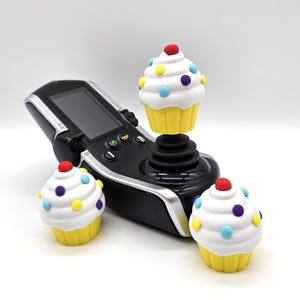 May include: A black electronic joystick controller with a screen and buttons, featuring a white cupcake topper with yellow base and colorful sprinkles. Two additional matching cupcake toppers are positioned in front of the controller.