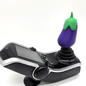 May include: A black and silver handheld gaming device with a purple and green aubergine-shaped joystick. The aubergine joystick is mounted on a black base. The device has a screen and several buttons.