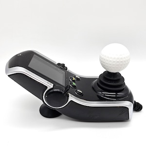 May include: Black and silver gaming controller with a white golf ball on top. The controller has a screen, buttons, and a joystick with a black rubber boot. The device is angled upwards.