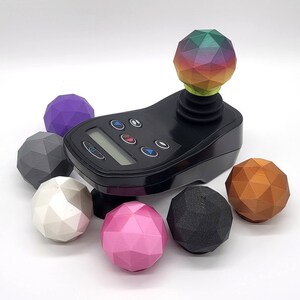 May include: A black joystick with a rainbow-coloured ball on top. There are six different coloured balls surrounding the joystick, including purple, grey, white, pink, black, and copper.