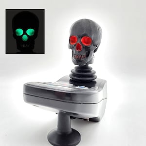 May include: A black skull with red eyes is mounted on a black joystick base. The skull glows green in the dark.
