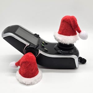 May include: A black gaming controller with two red and white Santa hats on the top. The controller has a screen and buttons.