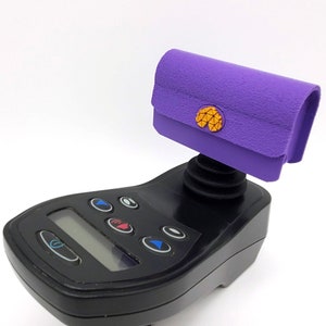 May include: A purple clutch with a gold emblem is mounted on a black electronic device. The clutch has a textured surface and a curved top. The device has a screen and several buttons.