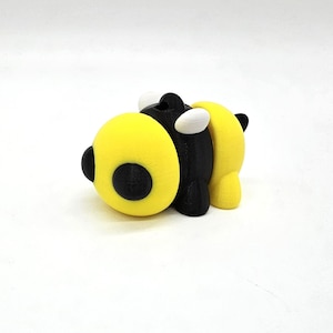 May include: A 3D-printed figurine of a stylized bee, featuring a yellow body with a black circle and black stripes. The bee has white wings and a black head. The figurine is set against a white background.