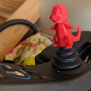 May include: A red, low-poly, 3D printed figurine of a Pokemon character, possibly Charmander, is mounted on a black joystick.