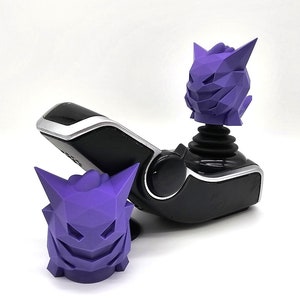 Peut inclure: Un joystick noir avec un personnage violet de forme géométrique sur le dessus. Le personnage est également disponible en figurine séparée, plus petite.