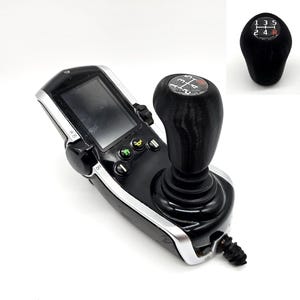 May include: A black joystick controller with a gear shift knob, featuring a screen and buttons. The gear shift knob has a 5-speed pattern. The controller has a silver trim and a black cable.