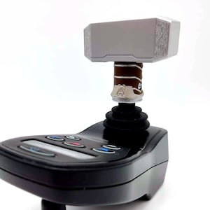 May include: A black joystick controller with a silver and brown hammer-shaped top. The hammer has a textured design on the sides and is attached to a brown and silver handle. The controller has several colored buttons.