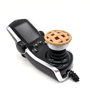 Peut inclure: Une manette de jeu noire et argentée avec une petite tartelette sur le dessus. La tarte a une croûte en treillis et une garniture rouge. La manette possède un écran et plusieurs boutons.