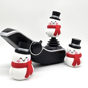 May include: A black and silver joystick with three small snowman figurines. The snowmen have black top hats, red scarves, and smiling faces. The joystick is on a white background.