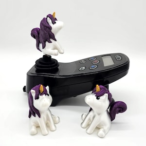 Könnte beinhalten: Drei weiße Einhorn-Figuren mit lila Mähnen und goldenen Hörnern. Die Einhörner sitzen auf einem schwarzen Joystick-Sockel.