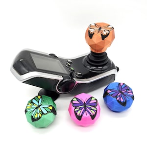 May include: A black mobility device joystick with a screen and buttons, topped with an orange geometric butterfly. Three additional butterfly-topped joysticks in green, pink, and blue are also present, each with a different butterfly design.