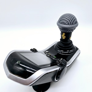 May include: A black and silver gaming controller with a microphone attached to the top. The microphone is gray and has a black and gold musical note design.