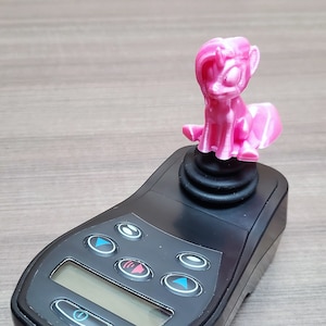 Könnte beinhalten: Eine rosafarbene Einhornfigur sitzt auf einem schwarzen Joystick mit Knöpfen und einem digitalen Display.