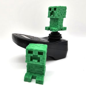 Peut inclure: Deux figurines Creeper Minecraft vertes. L'une est sur un joystick noir, l'autre est debout sur une surface blanche.