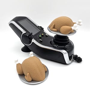 Peut inclure: Un contrôleur de joystick noir avec un écran et deux figurines de dindes rôties brunes sur le dessus.