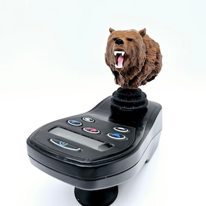 May include: A brown bear figurine with an open mouth is mounted on top of a black joystick with multiple buttons. The joystick has a digital display.