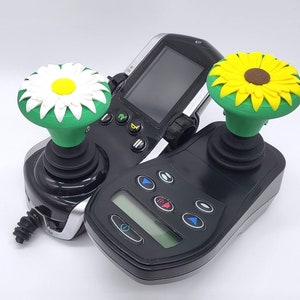 May include: Two black joysticks with green and white flower-shaped tops. The joysticks have multiple buttons and a screen on the side.