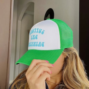 May include: A white and green trucker hat with a blue and white text design that reads "Drinking my day away, surrounded by you".