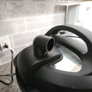 May include: Close-up of a black electric pressure cooker with a black plastic steam release valve. The valve is shaped like a 90-degree elbow.