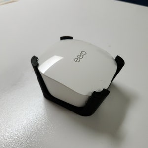 May include: A white eero router is sitting in a black plastic stand. The router has the word "eero" printed on the top.