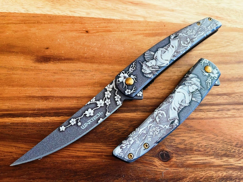 May include: A gray folding knife with a detailed geisha design and cherry blossom accents. The knife features the text "WARTECH" and gold-colored hardware. The blade is open, and another closed knife is next to it.