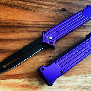 Könnte beinhalten: Zwei lila-schwarze Messer werden auf einer Holzoberfläche gezeigt. Die Messer haben ein schlankes Design mit einem lila Griff und einer schwarzen Klinge. Ein Messer ist geöffnet und zeigt die volle Klinge, während das andere geschlossen ist.