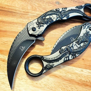 May include: A black folding knife with a dragon design on the handle. The knife has a curved blade and a ring on the handle.