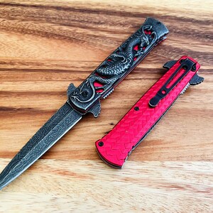 May include: Two pocket knives with a dragon design on a wooden surface. One knife has a red handle and a black blade. The other knife has a black handle and a silver blade.