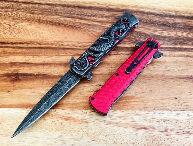 May include: Two pocket knives with a dragon design on a wooden surface. One knife has a red handle and a black blade. The other knife has a black handle and a silver blade.