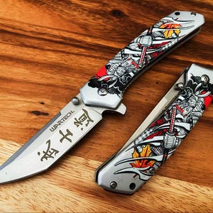 May include: A folding pocket knife with a silver blade featuring the text WARTECH and Japanese characters, alongside a closed folding knife. Both handles display a detailed graphic of a samurai in armour with red, black, and yellow maple leaf accents.