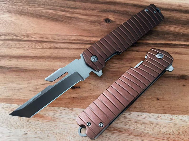 May include: Two brown folding knives with silver blades are shown on a wooden surface. The knives have a textured handle design and a small loop at the base. One knife is open, the other closed.