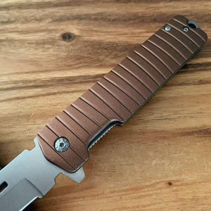 May include: A brown and silver folding knife is shown in close-up. The handle has a textured, ridged design. The blade is silver and partially open. The knife rests on a wooden surface.