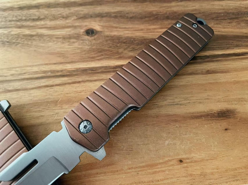 May include: A brown and silver folding knife is shown in close-up. The handle has a textured, ridged design. The blade is silver and partially open. The knife rests on a wooden surface.
