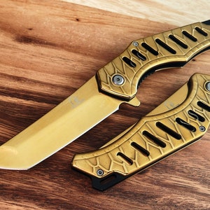 Puede incluir: Un cuchillo de bolsillo de color dorado con un mango estampado y una hoja afilada. El cuchillo está abierto y la hoja está extendida.