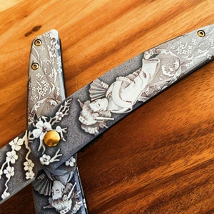 May include: A pair of open scissors with a detailed Japanese-inspired design. The blades feature a gray background with white floral patterns, geisha figures, and gold accents. The scissors are set against a wooden surface.