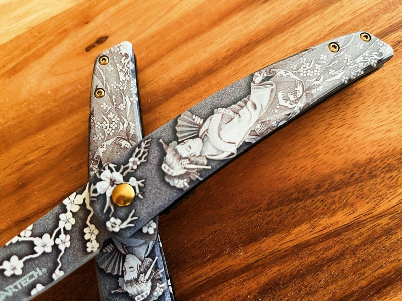 May include: A pair of open scissors with a detailed Japanese-inspired design. The blades feature a gray background with white floral patterns, geisha figures, and gold accents. The scissors are set against a wooden surface.