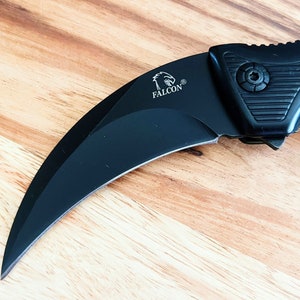 Pu&ograve; includere: Coltello pieghevole nero con lama curva e manico nero. Il manico ha una presa testurizzata e la lama ha la parola "FALCON" incisa.