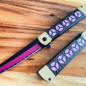Könnte beinhalten: Ein schwarzes Klappmesser mit einem rosa und goldenen Griff. Die Klinge ist teilweise geöffnet und hat eine gezackte Kante.