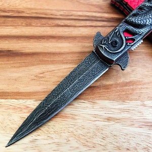 May include: A silver folding knife with a dragon design on the handle. The blade is partially open and has a detailed pattern.