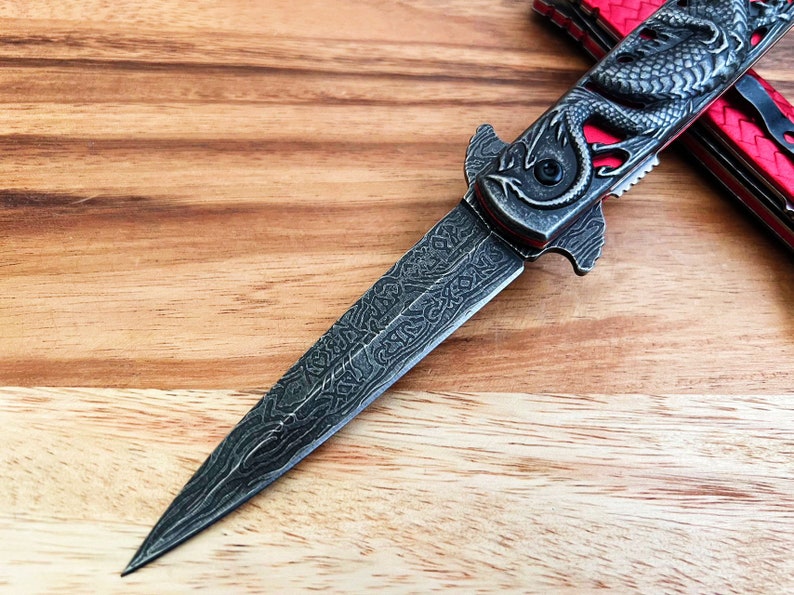 May include: A silver folding knife with a dragon design on the handle. The blade is partially open and has a detailed pattern.