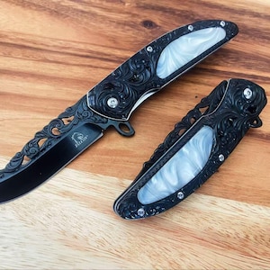 Op de afbeelding: Twee inklapbare messen met zwarte handgrepen voorzien van ingewikkelde krulgravures en witte parelmoer inlays. Eén mes is volledig uitgeklapt en toont een zwart lemmet met de tekst Falcon, terwijl het andere mes gesloten is.