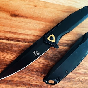 May include: A black folding knife with a black handle and a gold accent. The knife is open and the blade is visible. The knife has the word "FALCON" engraved on the blade.