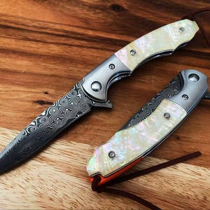Könnte beinhalten: Ein Klappmesser mit einer Damaststahlklinge mit Wirbelmuster, einem Griff mit schillernden weißen Perlmutt-Einlagen und einem braunen Lederband. Ein zweites identisches geschlossenes Messer liegt neben der geöffneten Klinge.