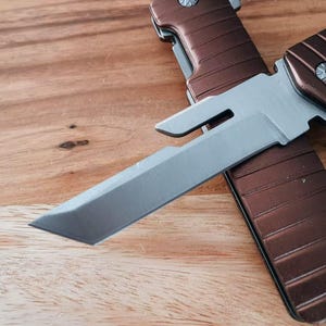 May include: A brown and silver folding knife is shown in close-up. The silver blade is partially open, and the handle is brown. The knife rests on a wooden surface.