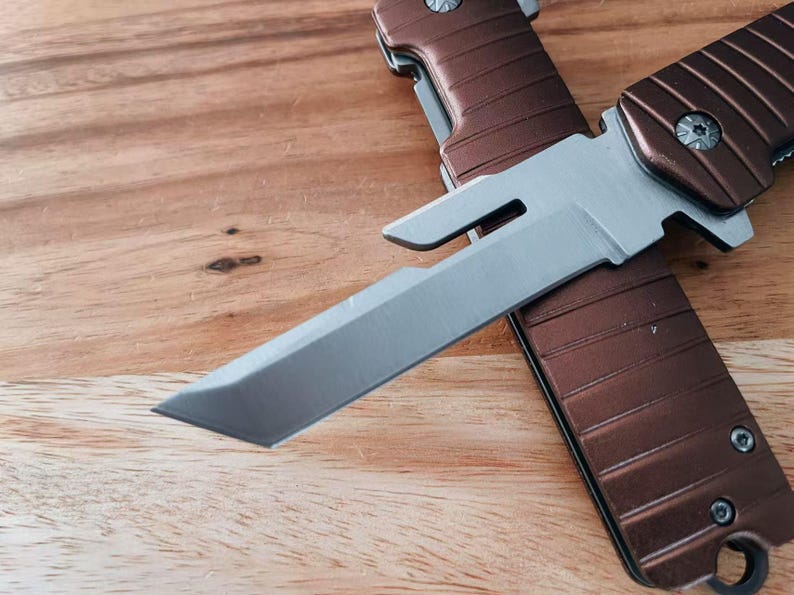 May include: A brown and silver folding knife is shown in close-up. The silver blade is partially open, and the handle is brown. The knife rests on a wooden surface.