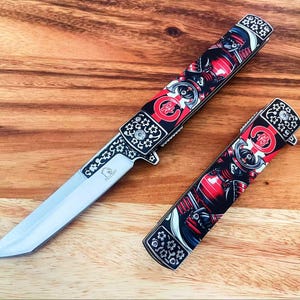May include: A folding knife with a silver blade and a handle featuring a red and black samurai graphic, alongside a matching closed folding knife handle. Both handles display white cherry blossom patterns and a red circular emblem with Japanese text.
