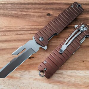 May include: Two brown and silver folding knives on a wooden surface. The knives have a textured handle design and a silver blade. One knife is open, revealing the blade, the other is closed. The wooden surface is a light brown color.