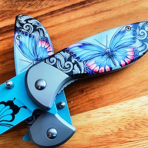 May include: A blue and silver pocket knife with a butterfly design. The knife has a black handle and a silver blade. The butterfly design is on both sides of the knife.