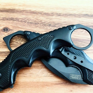 Pu&ograve; includere: Coltello tattico nero con manico ad anello e lama nera. Il coltello ha un'impugnatura testurizzata e un blocco di sicurezza. La lama &egrave; parzialmente aperta e il coltello &egrave; appoggiato su una superficie di legno.